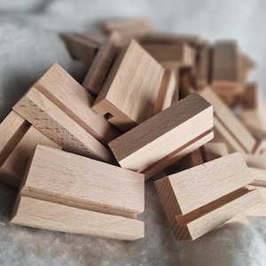 Op de afbeelding: Een stapel kleine, rechthoekige houten blokken met elk een gleuf erin. De blokken hebben een lichte, natuurlijke houtkleur en lijken onafgewerkt te zijn. De blokken zijn waarschijnlijk bedoeld om items zoals telefoons of kleine borden vast te houden.