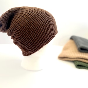 Peut inclure: Un bonnet en tricot marron exposé sur une tête de mannequin blanche. Le chapeau a un motif texturé en forme de gaufre. En arrière-plan, trois autres bonnets beiges, gris et vert olive sont légèrement flous.