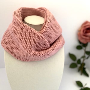 May include: A dusty rose-colored knitted infinity scarf displayed on a mannequin head. The scarf is made of a thick, textured yarn and is draped in a figure-eight style. A soft pink rose is slightly out of focus in the background, adding a touch of elegance.
