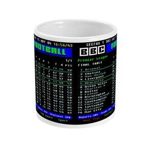 K&ouml;nnte beinhalten: Wei&szlig;er Keramikbecher mit einem Retro-Fu&szlig;balltabellen-Grafik von BBC Ceefax. Das Design zeigt Team-Tabellen, Daten und das Wort "FOOTBALL". Ein einzigartiges Geschenk f&uuml;r Sportfans.
