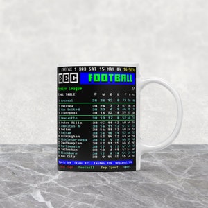 K&ouml;nnte beinhalten: Wei&szlig;e Keramik-Tasse mit einem Retro-BBC-Fu&szlig;ball-Grafik, die eine Abschlusstabelle der Premier League zeigt. Die Tasse zeigt die Team-Wertungen, einschlie&szlig;lich Arsenal, Chelsea und Manchester United, mit Datum und Uhrzeit.