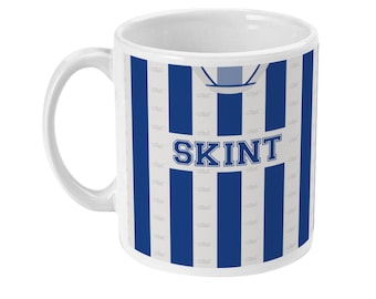 May include: White ceramic mug with blue and white vertical stripes. The word "SKINT" is printed in blue block letters on the front of the mug. The mug has a white handle and a white interior.