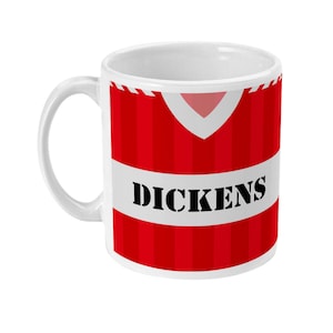 Könnte beinhalten: Weiße Keramik-Tasse mit rot-weißem Streifendesign, mit dem Namen "DICKENS" in fetten schwarzen Buchstaben. Die Tasse hat einen Henkel und ein Design, das an ein Trikot erinnert.