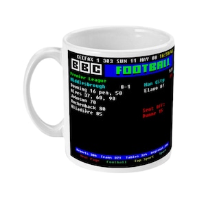 May include: White ceramic mug featuring a retro BBC Football graphic with game scores and team names. The mug has a curved handle and a black background with green and white text. The mug is a standard size.