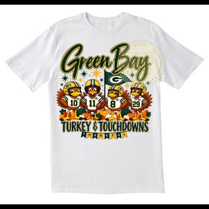 Könnte beinhalten: Ein weißes Kurzarm-T-Shirt mit einer Grafik von vier Truthähnen, die Footballhelme und Trikots mit den Nummern 10, 11, 8 und 29 tragen. Der Text lautet Green Bay, Turkey & Touchdowns und Thankful, umgeben von herbstlichen Kürbissen.