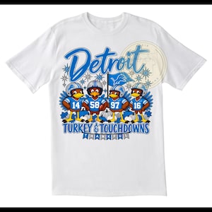 May include: A white t-shirt featuring a graphic of four cartoon turkeys wearing blue and silver football helmets and jerseys with the numbers 14, 58, 97, and 16. The text reads Detroit, Turkey & Touchdowns, and Thankful, with blue accents.