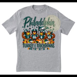 May include: A heather gray t-shirt featuring a graphic of four turkeys wearing football helmets and jerseys with the numbers 1, 26, 6, and 65. The text reads Philadelphia, TURKEY & TOUCHDOWNS, and THANKFUL, accompanied by autumn leaves and pumpkins.