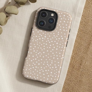 May include: A beige phone case with a white polka dot pattern. The phone case is lying on a cream-colored fabric, with a sprig of dried leaves and a burlap surface in the background. The phone case has a black camera module.