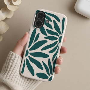 Puede incluir: Una funda de teléfono inteligente con un diseño floral en verde azulado sobre un fondo beige. La funda se sostiene en una mano, con unos auriculares inalámbricos blancos y una ramita de flores secas cerca. La funda tiene una cámara de doble lente.