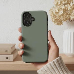 Könnte beinhalten: Eine salbeigrüne Handyhülle mit schwarzer Kameraobjektivabdeckung wird in einer Hand gehalten. Die Handyhülle ist glatt und matt. Der Hintergrund umfasst eine weiße Vase mit Trockenblumen und ein Holzregal mit Büchern.