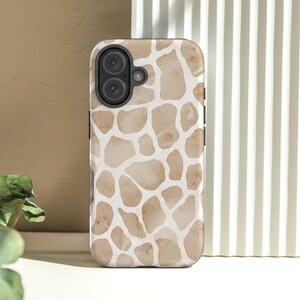 May include: A smartphone case with a giraffe pattern in shades of tan and cream. The case has a black camera module and buttons. The background includes a beige wall and a white striped surface.