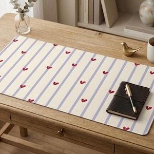Puede incluir: Una alfombrilla de escritorio rectangular con fondo color crema, rayas verticales azul claro y un patrón repetitivo de pequeños corazones rojos. La alfombrilla tiene esquinas redondeadas y una superficie lisa para el escritorio.