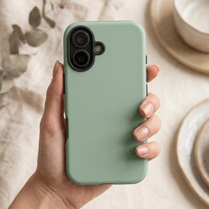Op de afbeelding: Een saliegroene smartphonehoes met een gladde, matte afwerking, voorzien van een zwarte ronde camera-uitsparing in de linkerbovenhoek die plaats biedt aan twee cameralenzen en een kleine ronde flitsopening aan de rechterkant van de module.
