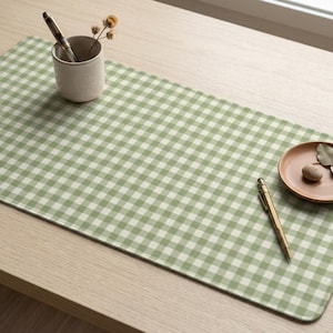 Può includere: Un tappetino da scrivania rettangolare con un motivo a quadretti vichy verde salvia e bianco. Il tappetino è decorato con una piccola tazza in ceramica beige contenente una penna e fiori secchi, una penna dorata e un piattino in terracotta.