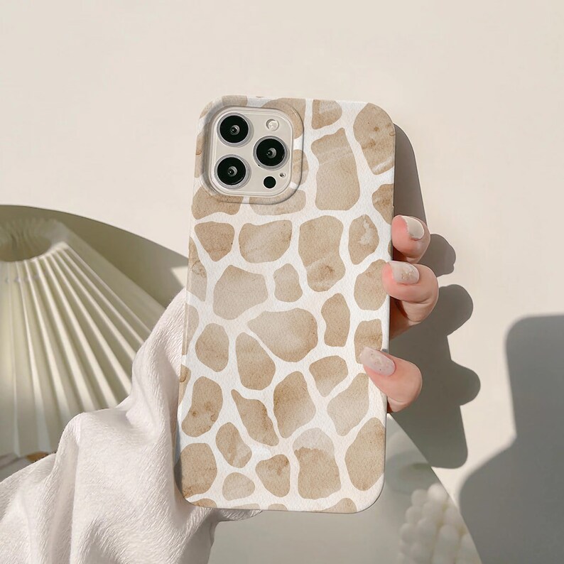 May include: A phone case featuring a giraffe print design in shades of tan and white. The case has a smooth finish and is designed to fit a smartphone. The case is held by a person.