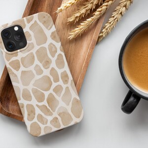 May include: A smartphone case with a giraffe print design in shades of tan and cream. The case is resting on a wooden tray with a cup of coffee and wheat stalks. The case has a camera and flash.