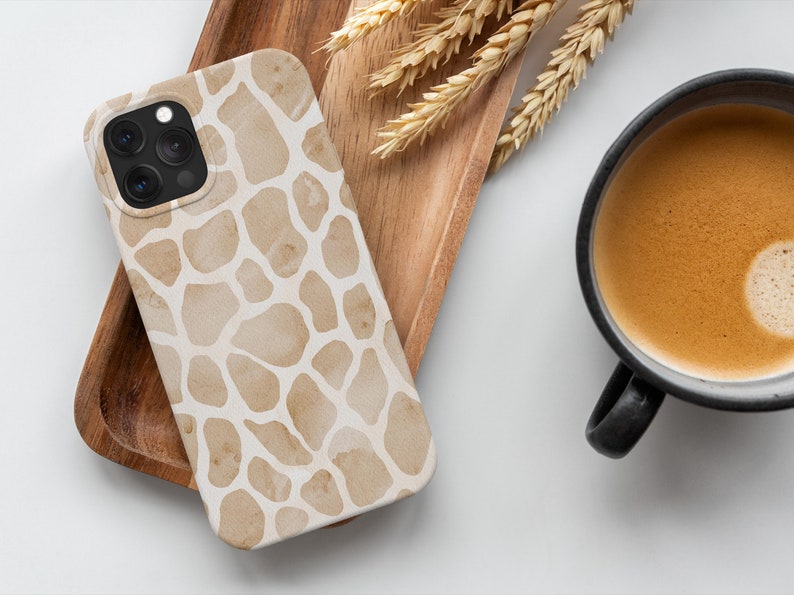 May include: A smartphone case with a giraffe print design in shades of tan and cream. The case is resting on a wooden tray with a cup of coffee and wheat stalks. The case has a camera and flash.