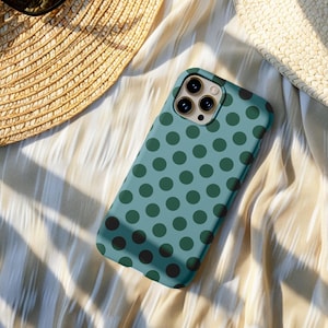 May include: A teal phone case featuring a pattern of dark green polka dots. The case has a camera cutout and is on striped fabric. A straw hat is partially visible in the background.