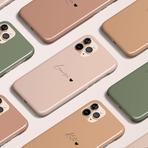 Puede incluir: Nueve fundas de teléfono diferentes con nombres personalizados y un pequeño diseño de corazón. Las fundas son de varios colores, incluyendo verde, rosa y marrón.