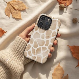 May include: A phone case with a giraffe print design in shades of beige and white. The case is held by a person. The background includes fall leaves and nuts.
