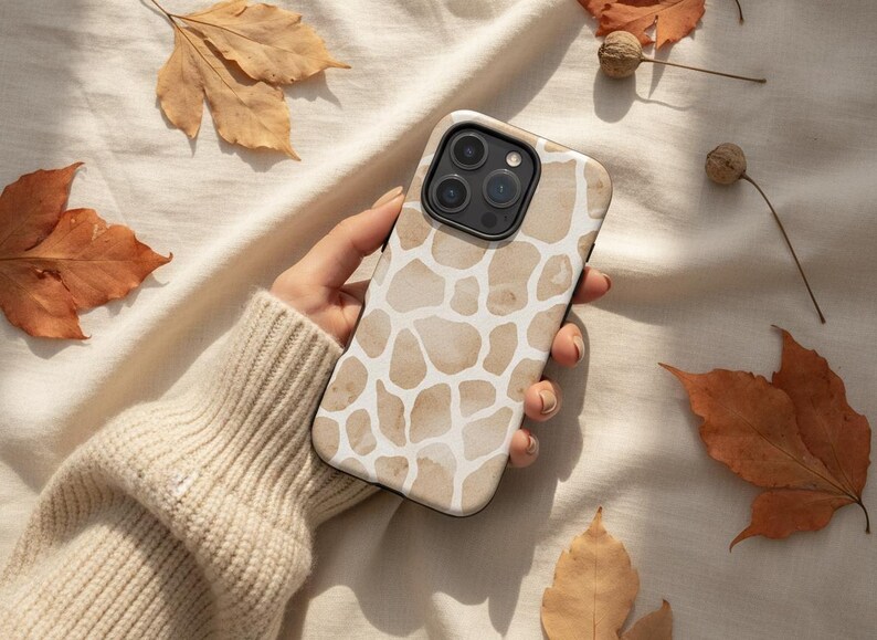 May include: A phone case with a giraffe print design in shades of beige and white. The case is held by a person. The background includes fall leaves and nuts.