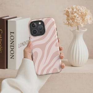 May include: A pink and white zebra-striped phone case. The case is held in a hand and features a camera cutout. The background includes books and a white vase with flowers.