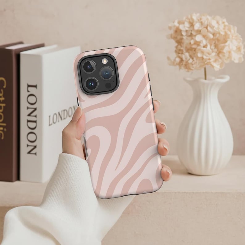 May include: A pink and white zebra-striped phone case. The case is held in a hand and features a camera cutout. The background includes books and a white vase with flowers.