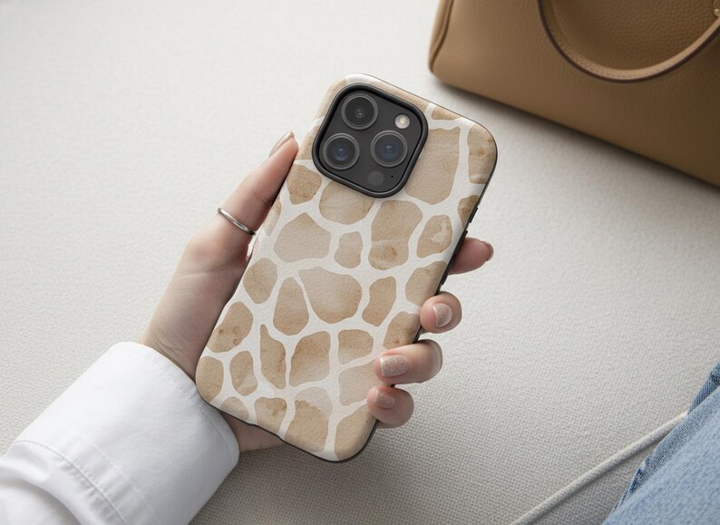 May include: A smartphone case with a giraffe print design. The case features a pattern of irregular brown shapes on a white background. The phone case has a black border and a camera module with three lenses.