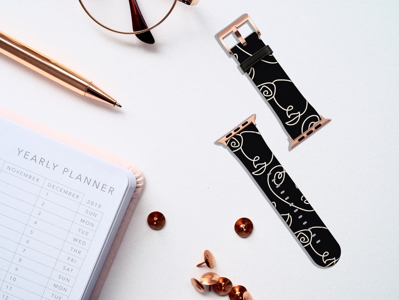 May include: Two black and white Apple Watch bands with rose gold hardware. The bands feature a repeating pattern of abstract faces.