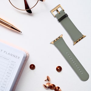 May include: Two gray leather Apple Watch bands with gold hardware. The bands are laid out on a white surface.