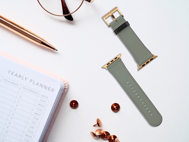 May include: Two gray leather Apple Watch bands with gold hardware. The bands are laid out on a white surface.