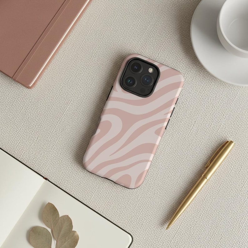 May include: A pink and white swirl pattern phone case. The case is on a light beige surface, next to a gold pen, a notebook, and a cup and saucer. The phone case has a black border and a camera lens.