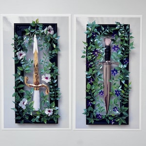 Puede incluir: Dos obras de arte enmarcadas presentan cada una una espada rodeada de hojas verdes y flores. Una espada tiene una empuñadura dorada y un mango blanco, la otra tiene una hoja plateada y un mango negro. Los marcos son negros.
