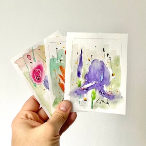 May include: Three watercolor floral paintings held in a hand. The paintings feature colorful flowers in shades of pink, purple, and orange, with green foliage and white accents. Each painting is on a white rectangular paper.