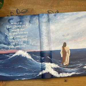 Puede incluir: Biblia abierta con una cubierta pintada que representa a Jesús caminando sobre el agua. La cubierta presenta una cita de Mateo 11:28, sobre un fondo de mar y cielo tormentosos. También son visibles las palabras "The Holy Bible".