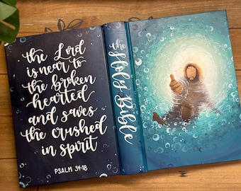 May include: A blue and black Holy Bible with a white inscription on the front cover that reads "the Lord is near to the broken hearted and saves the crushed in spirit Psalm 34:18". The back cover features a painting of a person in water with bubbles.