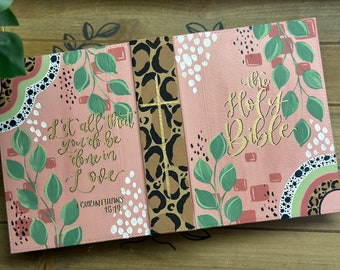 May include: A pink Bible cover with a leopard print spine and a gold cross. The front cover features a floral design with green leaves and white dots. The text "Let all that you do be done in love 1 Corinthians 16:14" is written in gold on the front cover. The back cover features the text "The Holy Bible" in gold.