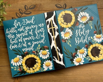 May include: A teal blue Bible cover with a cowhide pattern spine. The front cover features a white daisy and sunflower design with the text "for God hath not given us the spirit of fear but of power, & of love, & of a sound mind". The back cover features a similar design with the text "the Holy Bible".