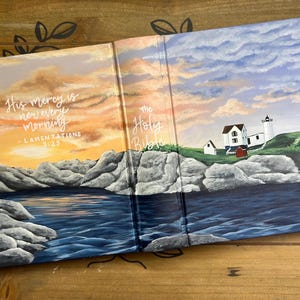 May include: Open Bible with a painted landscape scene. The left side depicts a sunset over water with the text "His mercy is new every morning - Lamentations 3:23." The right side shows a lighthouse and buildings under a cloudy sky.
