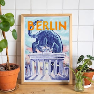 Könnte beinhalten: Gerahmter Kunstdruck mit einer blauen Illustration eines Tieres auf einem Gebäude und dem Wort "BERLIN" in Orange. Das Kunstwerk befindet sich in einem hellen Holzrahmen. Der Druck wird auf einer Holzoberfläche präsentiert.