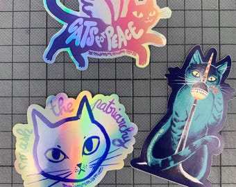 以下が含まれることがあります： 3つのホログラフィックステッカー。猫のイラストが特徴です。1つは翼のある猫で、「Cats for Peace」と書かれています。もう1つは「Smash the patriarchy」と書かれています。3つ目の猫はマイクのようなものを持っています。