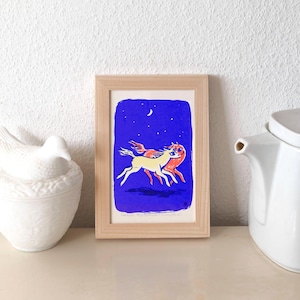 Könnte beinhalten: Ein gerahmter Kunstdruck mit zwei stilisierten Pferden, die über einen dunkelblauen Nachthimmel mit Mond und Sternen rennen. Die Pferde sind gelb und rot, vor blauem Hintergrund. Der Rahmen ist aus hellem Holz.