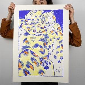 May include: A print depicting two leopards in a close embrace. The artwork uses yellow, blue, and orange hues, with the leopards' spotted patterns and the background in contrasting colours. The print is held up against a white background.