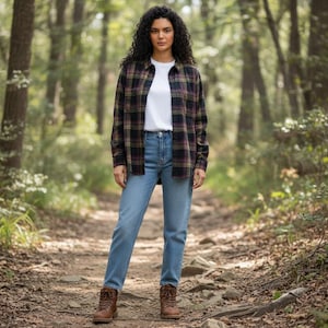 May include: A woman wearing a plaid flannel shirt in black, green, and purple over a white t-shirt, paired with light blue jeans and brown hiking boots. The background is a forest trail.