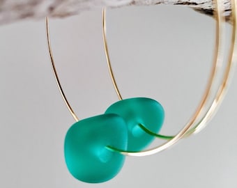 May include: Gold hoop earrings with teal green glass beads. The earrings are hanging from a piece of driftwood.