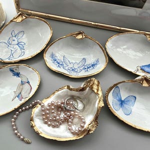 May include: Six decorative clam shells with white interiors and gold-painted edges. The shells feature illustrations of a blue rabbit, two blue birds, a rabbit in a blue jacket, a goose, a blue butterfly, and one shell holding a pearl necklace.