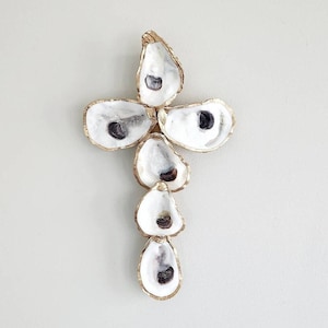 May include: A gold-painted oyster shell cross made of five oyster shells. The shells are arranged in a cross shape with the top shell being the largest and the bottom shell being the smallest.