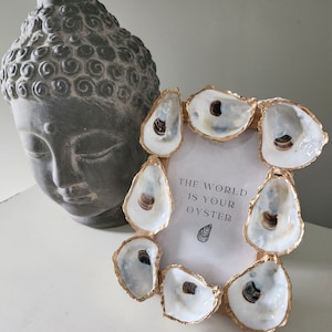 May include: A decorative photo frame adorned with oyster shells with gold trim. The frame holds a print with the text "THE WORLD IS YOUR OYSTER" and a small oyster illustration. A gray Buddha head statue is in the background.