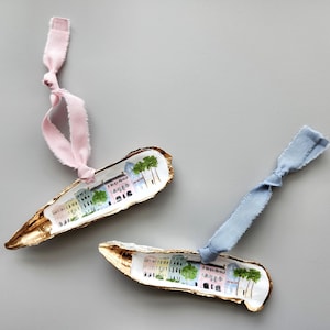 May include: Two oyster shell ornaments with gold leaf accents. Each shell is painted with a watercolor scene of colorful houses and palm trees. One ornament has a pink ribbon and the other has a blue ribbon.