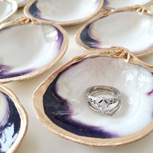 May include: A set of six white seashell ring dishes with gold trim and purple interiors. One dish has a silver ring with a diamond in the centre.